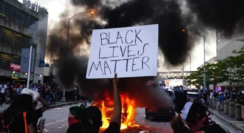2020년 5월 29일, 미국 애틀랜타 CNN 센터 밖에서 경찰차가 불타고 있는 가운데 한 남성이 '흑인 목숨도 소중하다(Black Lives Matter)' 팻말을 들고 있다. | Elijah Nouvelage/Getty Images